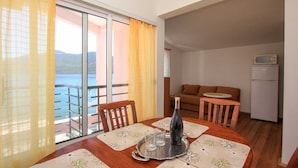 A dining area with a table set for two, a bottle, and glasses. There is a balcony with curtains and a view of the sea. A sofa and a small table are visible in the background. - Apartments Ereš Zuronja-One-Bedroom Apartment with Sea View 2 (Putnikovici)