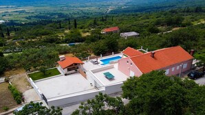 A residential area with a house featuring a swimming pool, a red-roofed terrace, and a view of a green landscape with hills.