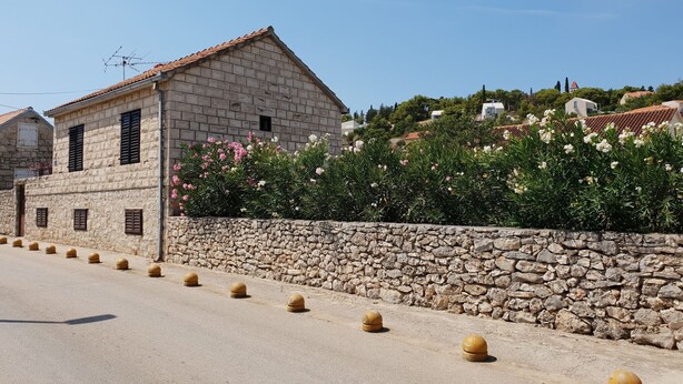 A stone building with a tiled roof, a stone wall, and a row of bollards along a paved road.