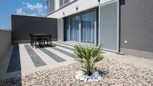 A modern outdoor patio with a dining table and chairs, a palm plant, and a building with large windows and shutters. - Apartments Dva Galeba - Standard One Bedroom Apartment with Terrace (101) (Bol)