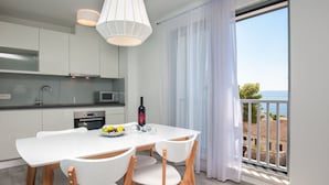 A modern kitchen with white cabinets, a dining table with chairs, a microwave, and a view of the sea through a balcony. - Apartments Dva Galeba - One Bedroom Apartment with Sea View (105) (Bol)