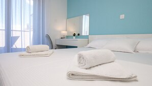Habitación de hotel con cama, dos toallas enrolladas, mesita de noche con lámpara, espejo y ventana con cortinas.