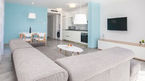 Una sala de estar moderna con un juego de sofá, una mesa de comedor con sillas, una zona de cocina con microondas y horno, y un televisor montado en la pared.