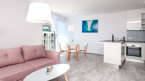 A modern living room with a pink sofa, a white coffee table, and a dining area with wooden chairs. There is a kitchen with white cabinets and a built-in oven. A large window with curtains is visible, and a blue abstract painting hangs on the wall. - Apartments Dva Galeba - Standard One Bedroom Apartment with Sea View (203) (Bol)