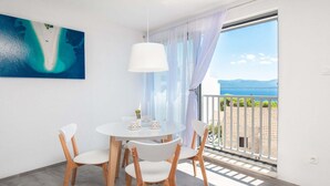 Ein Zimmer mit einem runden Tisch und vier Stühlen, einer weißen Pendelleuchte, einem Strandbild und einem Balkon mit Blick auf das Meer und die Berge.