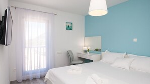 A hotel room with a bed, a desk with a chair, a TV mounted on the wall, and a window with sheer curtains. - Apartments Dva Galeba - One Bedroom Apartment with Sea View (206) (Bol)