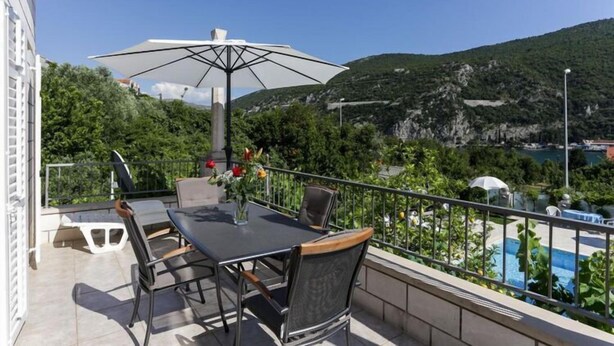 Ein Balkon mit Tisch und Stühlen, einem großen Sonnenschirm und Blick auf eine gebirgige Landschaft.