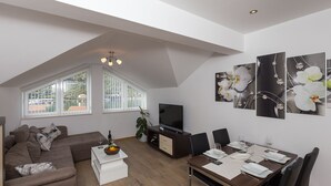 Ein modernes Wohnzimmer mit einem braunen Sofa, einem Esstisch für vier Personen, einem Fernseher und großen Fenstern mit Jalousien.