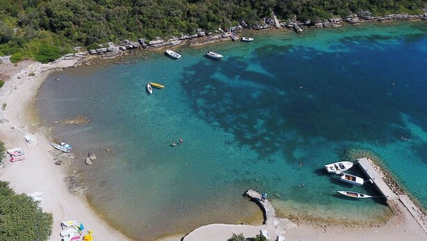 Una zona costera con barcos amarrados cerca de la orilla, una playa de arena y un promontorio boscoso al fondo.