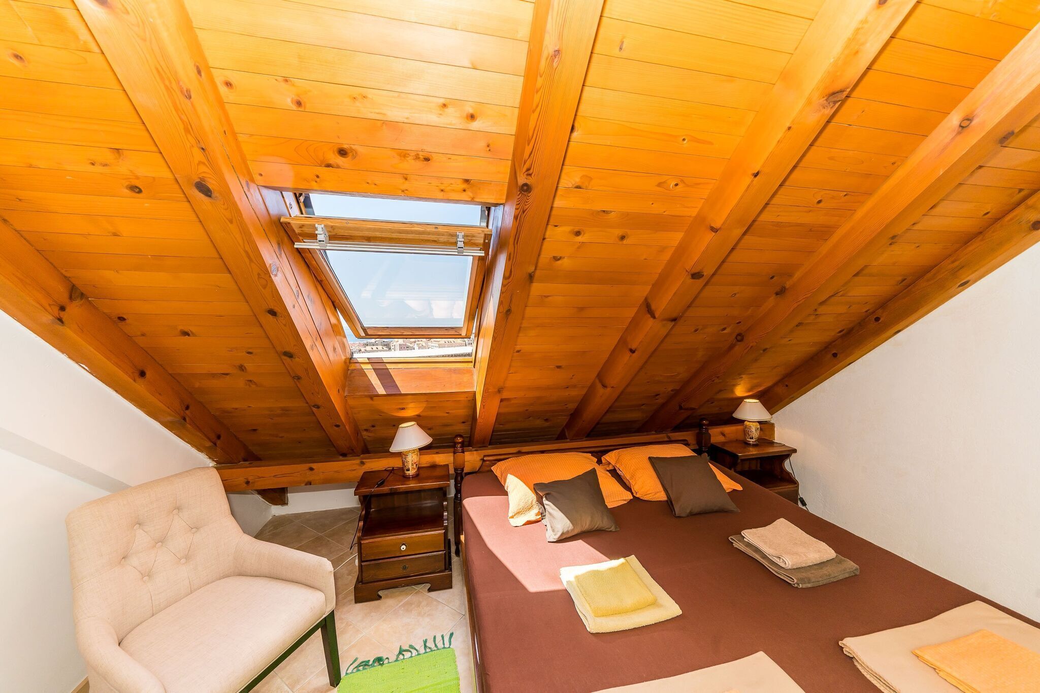 A cozy attic bedroom with a sloped ceiling, a skylight, a bed with pillows, a chair, and bedside tables with lamps.