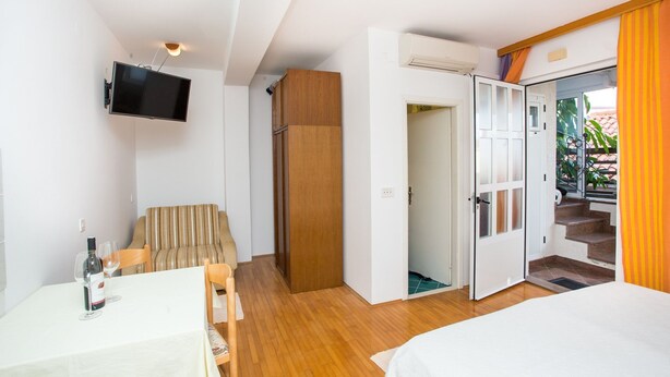 Un dormitorio con cama, televisor montado en la pared, mesa de comedor con sillas, sofá a rayas y armario de madera.