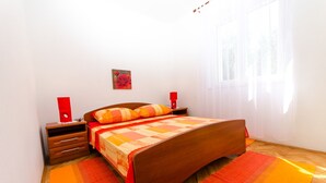 Un dormitorio con una cama de madera, dos mesitas de noche, un jarrón rojo en cada una, un cuadro de una rosa roja en la pared y una ventana con cortinas traslúcidas.
