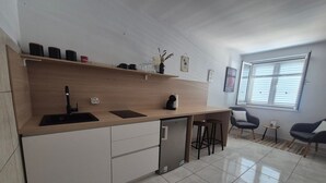 A modern kitchen with a dishwasher, a sink, and a countertop with a coffee maker. There are two bar stools and a small table near a window with blinds. - Apartments Matea - Studio with Sea View (2 Adults) - (Štikovica) (Stikovica)