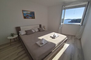 A bedroom with a bed, a window with a view, a small table with a plant, and a painting on the wall. - Apartments Matea - One-Bedroom Apartment with Sea View - (Štikovica) (Stikovica)