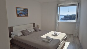 Un dormitorio con cama, almohadas, una mesita de noche, lámpara, un cuadro y ventana con vista al mar.