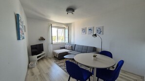 Una sala de estar moderna con un sofá gris, un televisor sobre un mueble blanco, una mesa de comedor redonda con sillas azules, y una lámpara de techo.