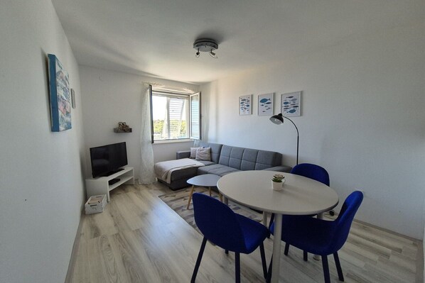 A modern living room with a grey sofa, a TV on a white stand, a round dining table with blue chairs, and a ceiling light. - Apartments Matea - One-Bedroom Apartment with Sea View - (Štikovica) (Stikovica)