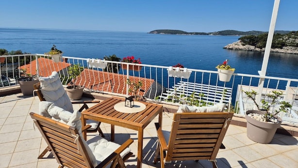Una terraza con mesa y sillas de madera, plantas en macetas y vista al mar.