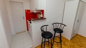 Une petite cuisine avec un mur recouvert de carreaux rouges, deux tabourets de bar noirs, une porte blanche et un plancher en bois.