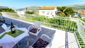 Une terrasse avec une table dressée pour deux personnes, une bouteille de vin et un verre. La terrasse offre une vue sur une ville, avec des bâtiments et des collines en arrière-plan.