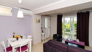 A modern living room with a white sofa, a dining table with a fruit bowl, and a view of a balcony with curtains.