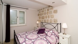 A bedroom with a bed, a nightstand, a lamp, a window with blinds, and a stone wall.