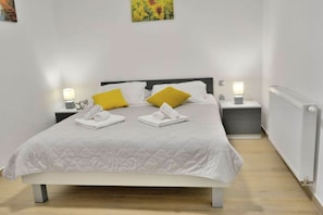 Una cama bien hecha con ropa blanca y dos almohadas, una amarilla y otra blanca, en una habitación con paredes blancas y un radiador.