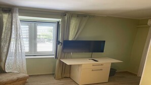 Ein kleines Zimmer mit Schreibtisch, Stuhl, Fernseher und einem Fenster mit Vorhängen.