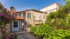 A stone house with a tiled roof, a satellite dish, and a garden with green plants and pink flowers.