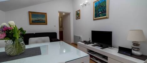 A modern living room with a glass table, white sofa, flat-screen TV, and artwork on the walls.