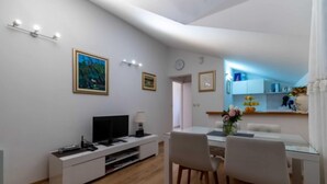 Un soggiorno moderno con televisione a schermo piatto, un mobile TV bianco, un tavolo da pranzo con sedie e una cucina visibile attraverso una porta aperta.