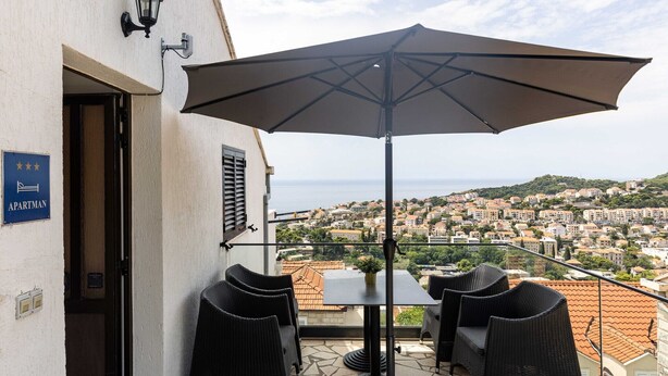 Un balcone esterno con un tavolo, sedie e un ombrellone grande. Un cartello indica che si tratta di un appartamento.