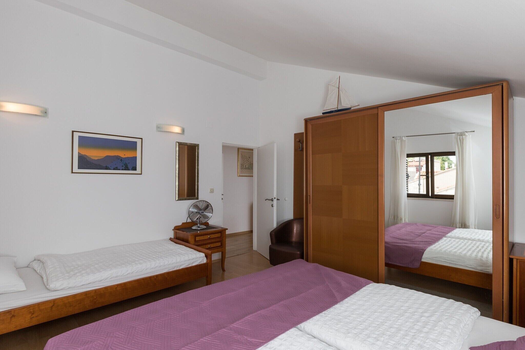 A room with two single beds, a wooden wardrobe, a desk with a chair, a standing fan, and a large mirror.