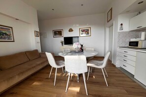 En moderne stue med et rundt spisebord, hvide stole, en brun sofa og et køkkenområde med en mikroovn.