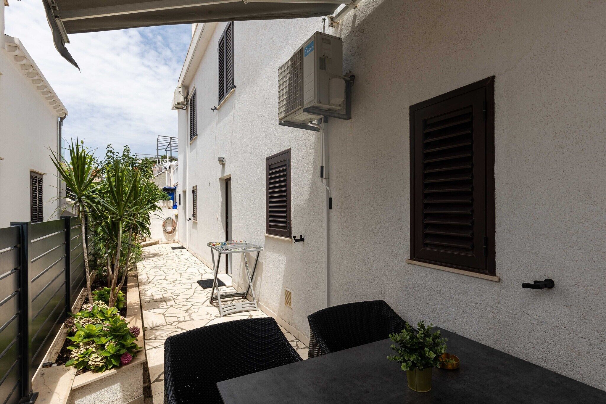 Un patio exterior estrecho con una mesa y sillas, un pequeño jardín y un edificio con persianas y un equipo de aire acondicionado.