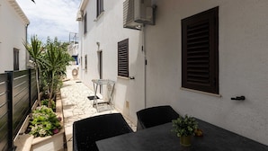 A narrow outdoor patio with a table and chairs, a small garden, and a building with shutters and an air conditioning unit.