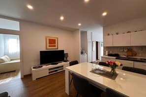 A modern living room with a flat-screen TV, a dining table with chairs, a sofa, and a kitchen area with cabinets and appliances.