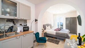 Ein kompaktes Schlafzimmer mit Bett, Nachttisch und Fernseher. Angrenzend befindet sich eine Küchenzeile mit Mikrowelle, Wasserkocher und Spüle.