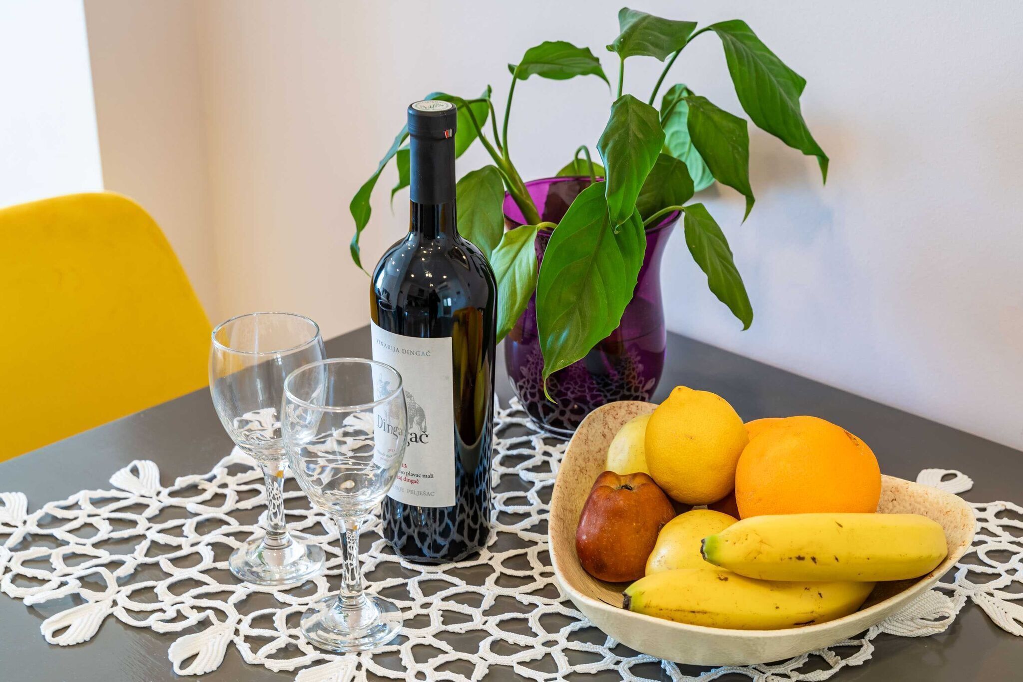 Une table avec une bouteille de vin, deux verres, un saladier de fruits et une plante en pot.