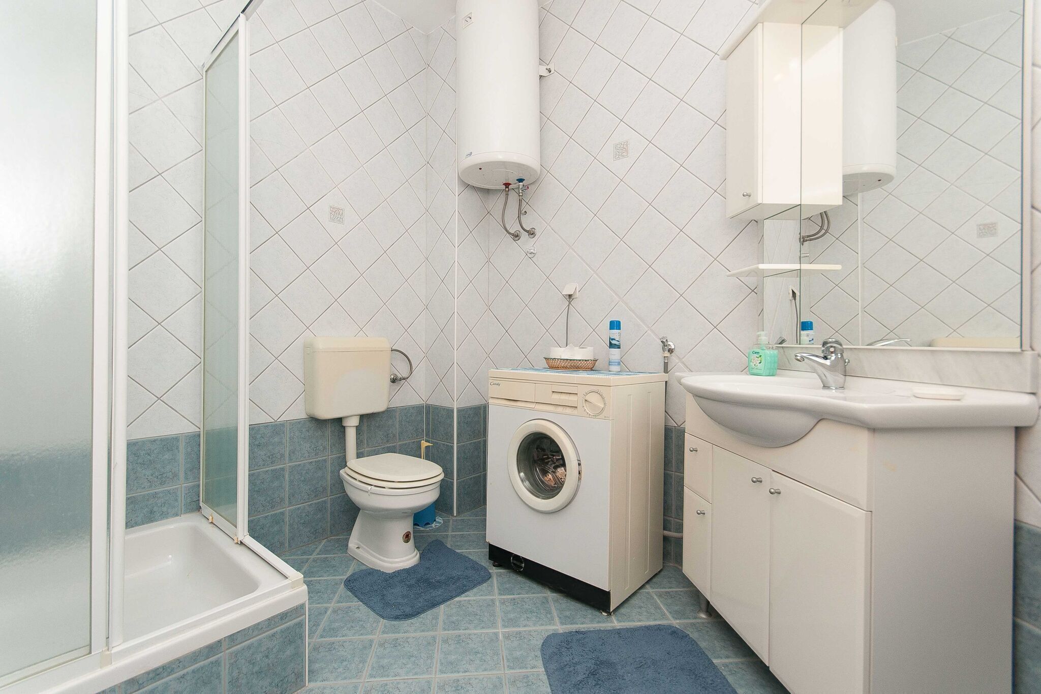 A bathroom with a shower, toilet, washing machine, sink, and tiled walls.