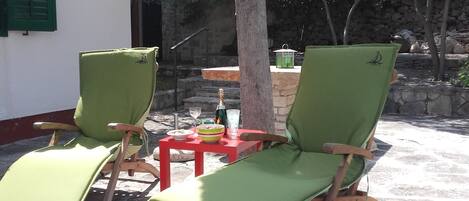 Two green cushioned lounge chairs with wooden frames, a small red table with a bottle and a bowl, and a stone wall in the background.