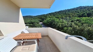 Ein Balkon mit weißem Stuhl, einem Holztisch und Blick auf einen bewaldeten Hang.