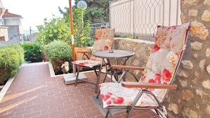 En udendørs terrasse med flisegulv, to metalstole med blomstermønstrede hynder, et rundt bord og en hvid parasol.