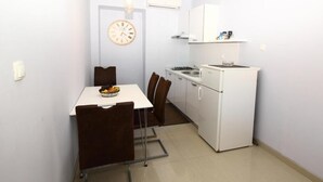 Cocina compacta con refrigerador, estufa y microondas blancos. Hay una mesa de comedor con dos sillas y un reloj en la pared.