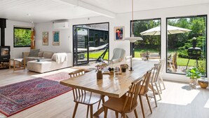 A spacious living area with a wooden dining table, comfortable seating, a large window with a view of the garden, and a built-in fireplace.
