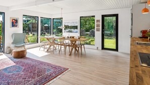 A spacious dining area with a large table and chairs, large windows offering views of a garden, and a modern kitchen with a wooden countertop.