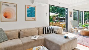 A modern living room with a beige sofa, a coffee table, and a bookshelf. There are two framed artworks on the wall and a large window offering a view of the outdoors.
