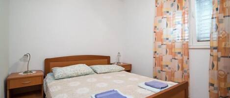 Een slaapkamer met een houten bed, twee nachtkastjes, een raam met gordijnen met een patroon, en een effen muur.