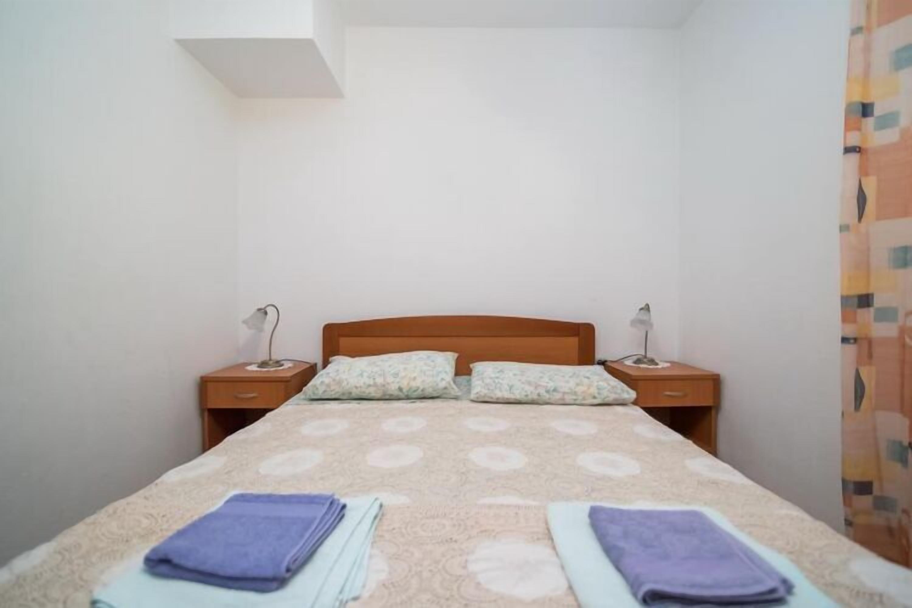 A single bed with a patterned bedspread, two pillows, two bedside tables with lamps, and a window with a curtain.