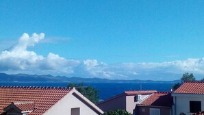A coastal residential area with red-tiled roofs and a clear view of the sea and distant mountains.